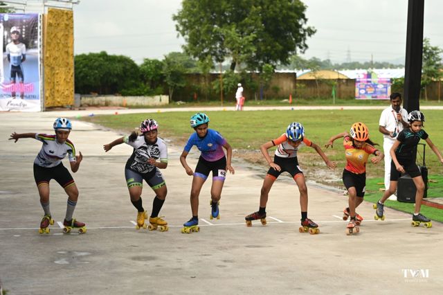 Six quad skaters crouched at the start line while an official speaks into a microphone